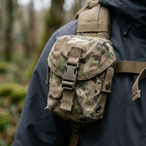 Close-up of a weathered MTP camouflage utility pouch attached to a tactical rucksack strap against a soft woodland background.