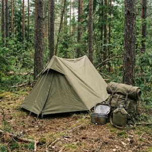 Camping site in a forest with an olive green tent, a vintage military rucksack, and a classic metal mess kit for bushcraft.