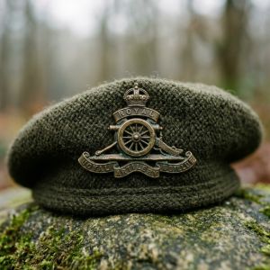 Macro shot of an authentic brass British Army cap badge with a rich patina, pinned to an olive drab wool beret on mossy stone.
