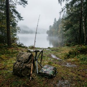 A fishing rod standing next to a vintage rucksack and a folded camo tarp by a calm, misty lake at sunrise.