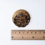 A measurement photo of the Royal Air Force cap badge on its backing plate next to a metric ruler.