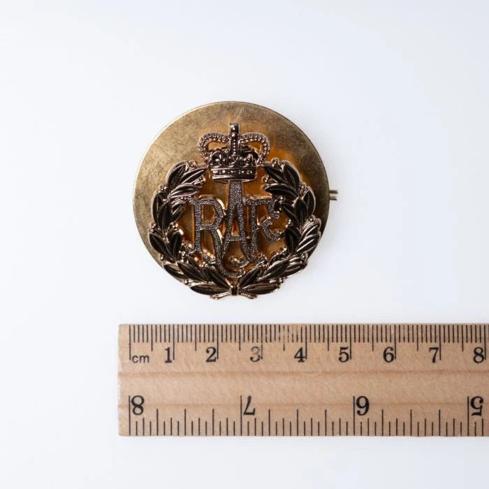 A measurement photo of the Royal Air Force cap badge on its backing plate next to a metric ruler.