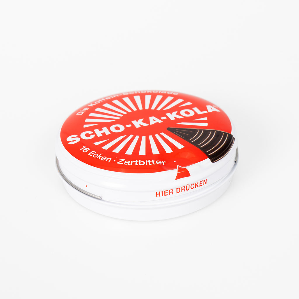 Side profile of a closed Scho-Ka-Kola metal tin, highlighting the press-to-open lid mechanism labeled Hier Drücken.