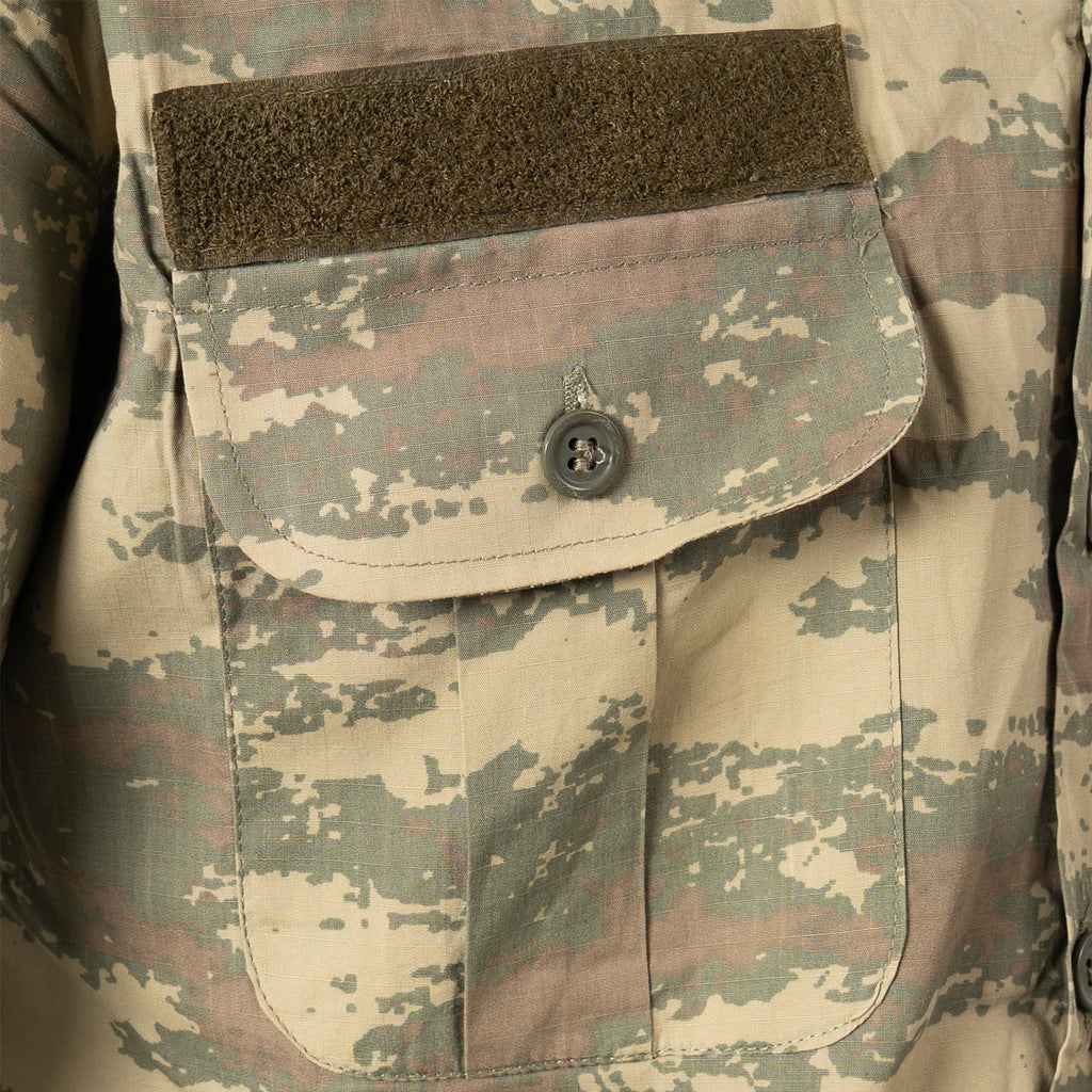 Close-up of the military field shirt chest pocket detail, showing the buttoned flap closure and a green Velcro strip for identification or insignia attachment.