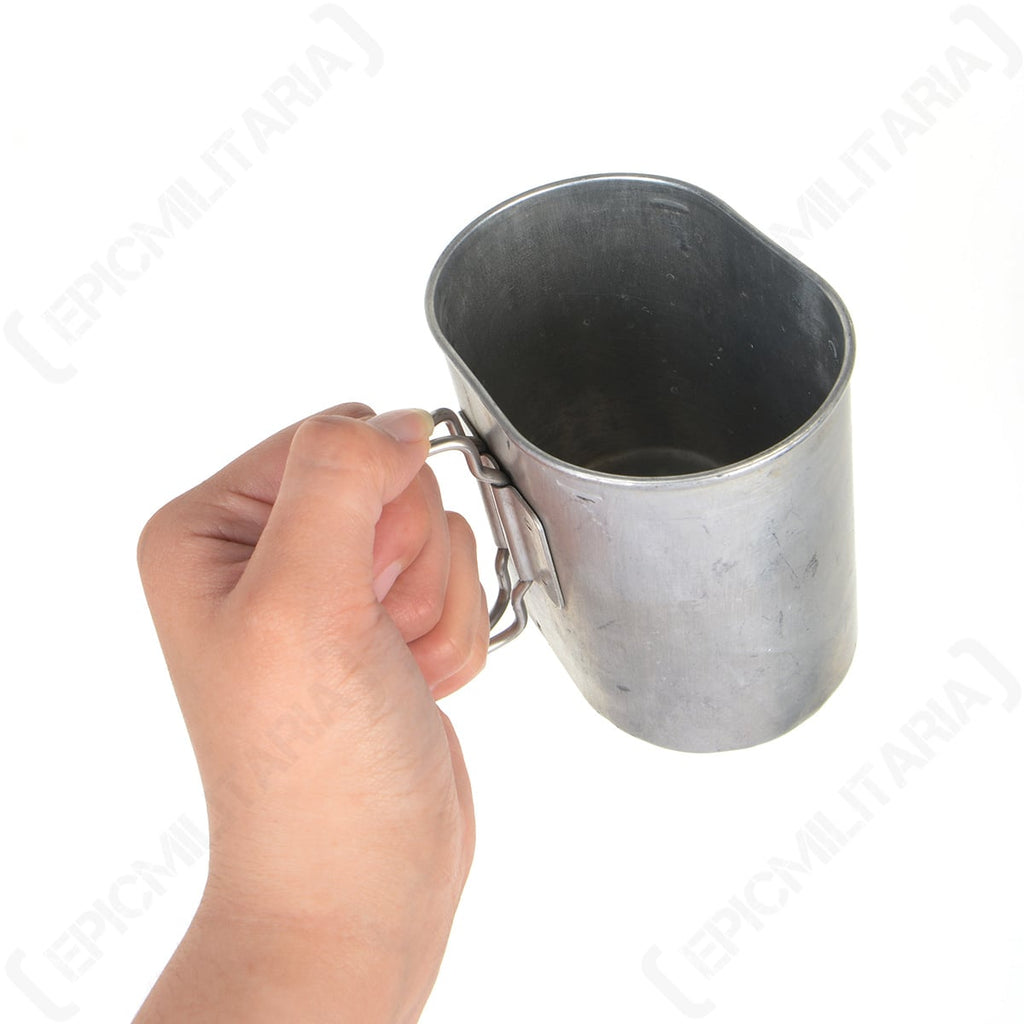 Close-up of a hand holding the oval aluminium canteen cup by its unfolded wire handle, demonstrating its utility as a camping mug.