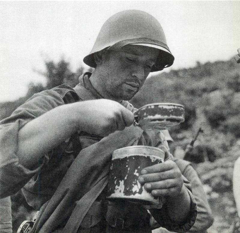 Black and white historical photograph of a soldier using a traditional metal mess tin in the field.