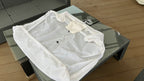 A plain white protective utility sheet laid flat on a table, featuring an elasticated hem and integrated storage pocket.