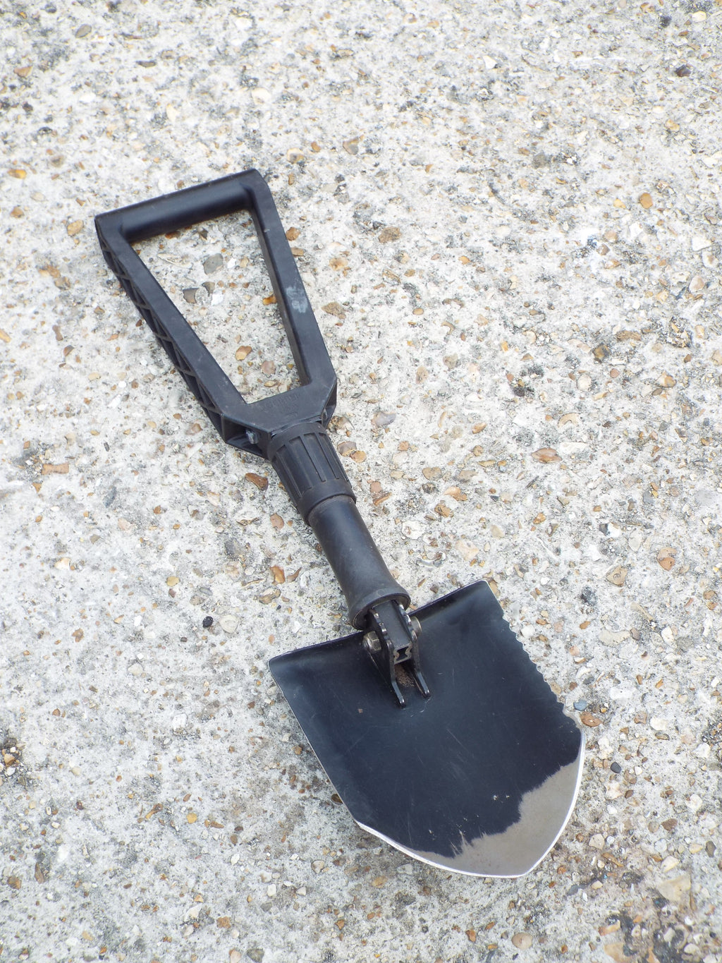 US Army Gerber E-Tool folding shovel in full shovel configuration with black steel blade and nylon handle on textured ground.