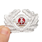 East German Army enlisted man visor cap badge held in hand to demonstrate the compact four by six centimetre scale