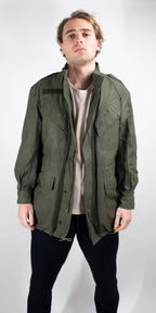 Front view of a man wearing a heavy duty cotton canvas Belgian military surplus field jacket in olive green with multiple utility pockets.