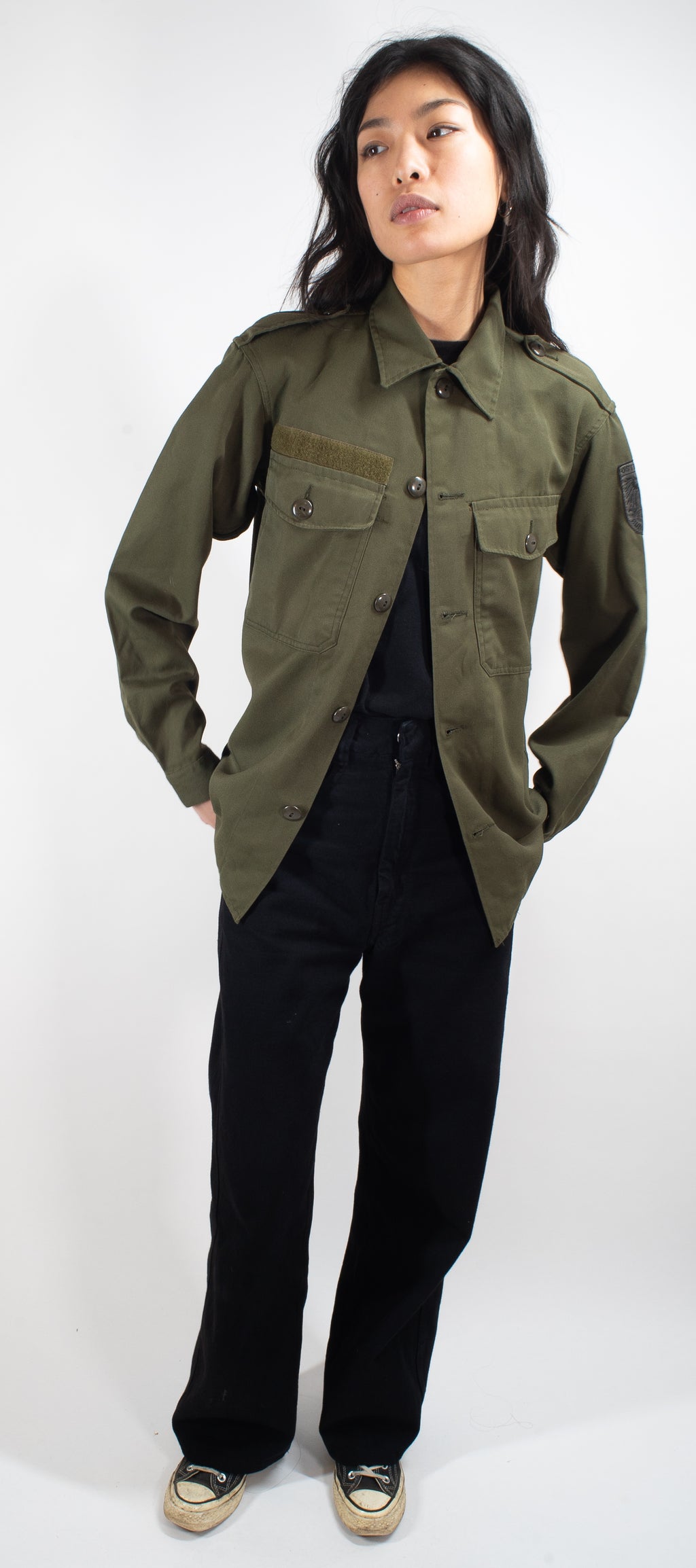 Full-body front view of a woman modeling the Austrian olive combat shirt with hands in pockets, showing length and classic collar.