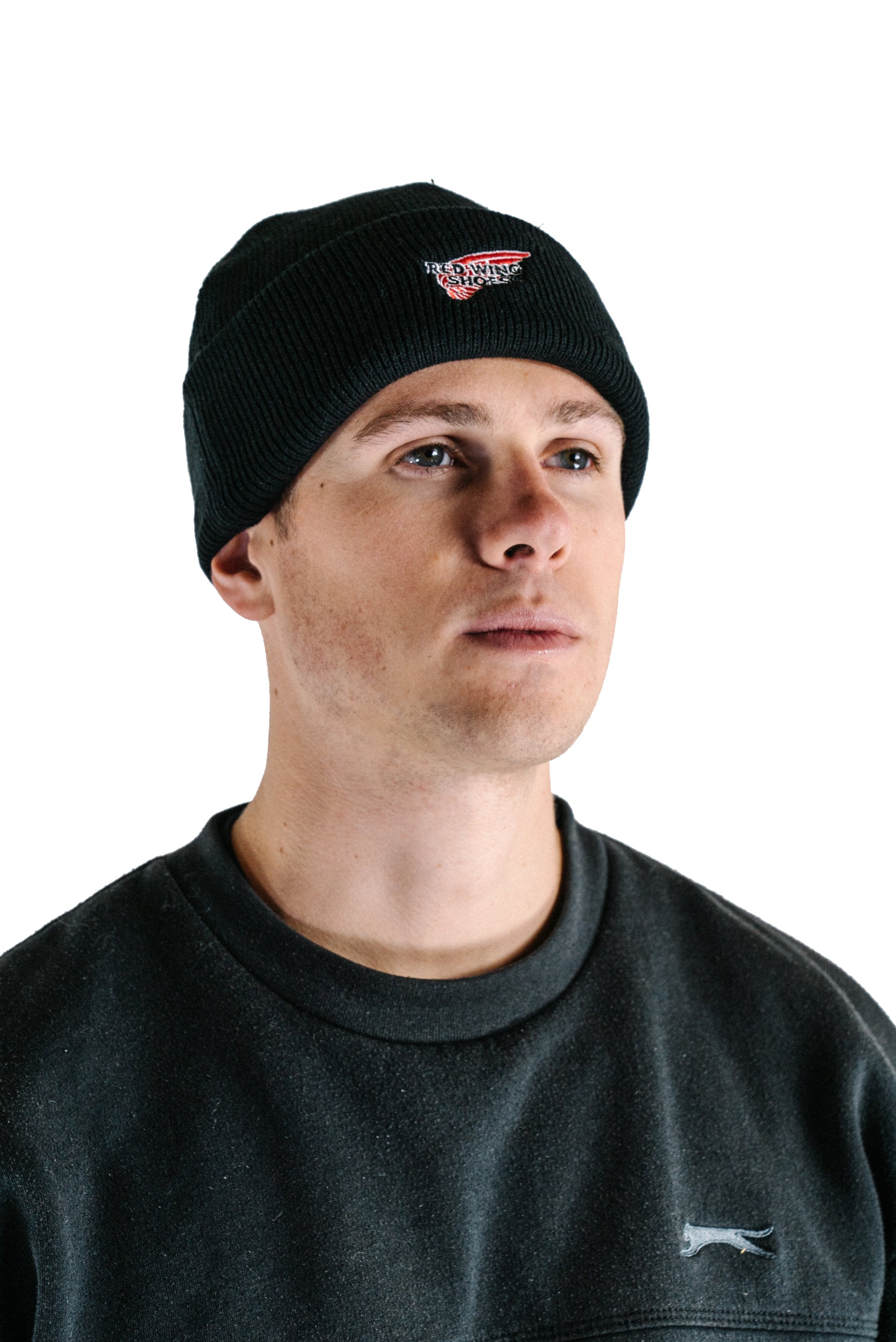 Model wearing a black 100% acrylic Red Wing Shoes beanie with a red and white embroidered wing logo on the front fold.