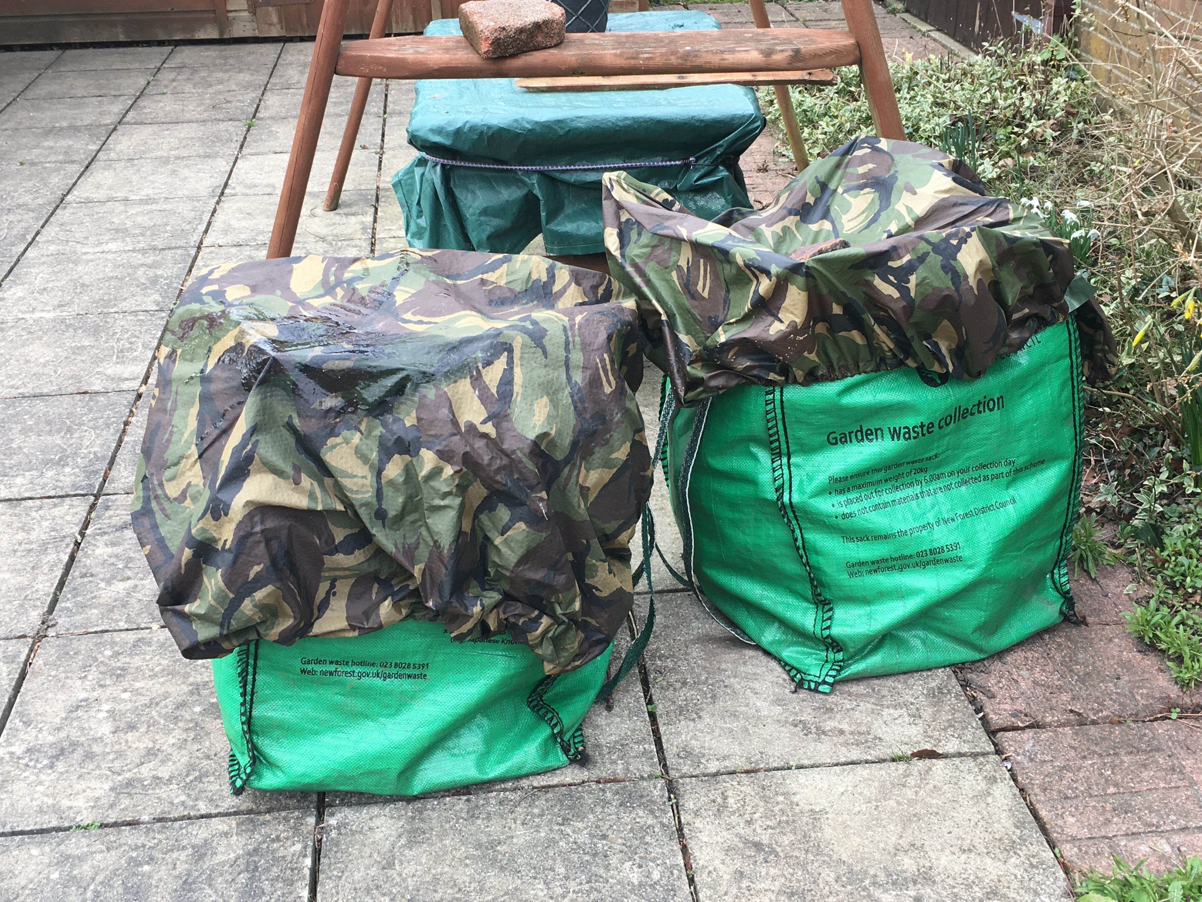 Two green garden waste bags covered by waterproof Dutch Army woodland camouflage sheets on a stone patio.