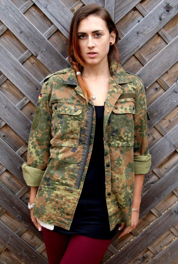Woman wearing an authentic vintage German flecktarn camouflage field shirt with sleeves rolled up over a dark outfit.