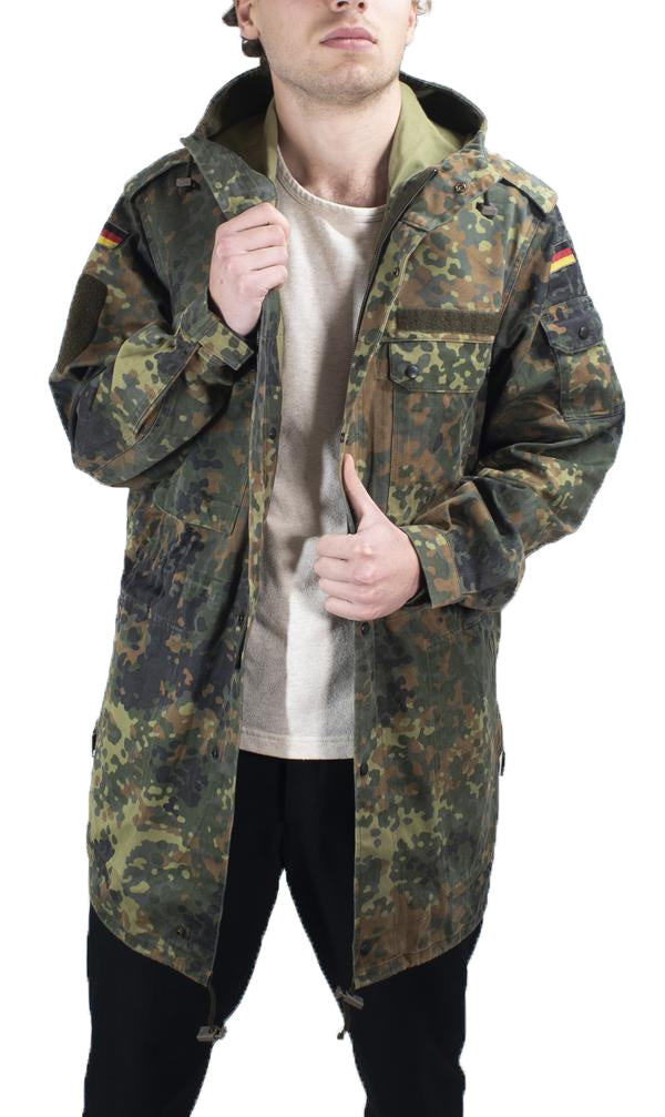 Man wearing a Flecktarn camouflage field jacket, displaying the chest cargo pockets and hood drawcords.